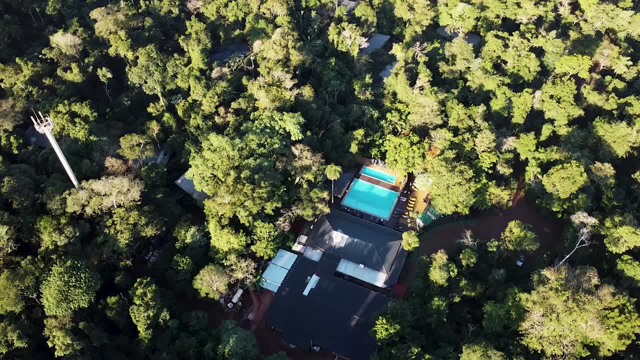 Aerial rotating drone shot reveals a jungle resort complex nestled in dense tropical vegetation with a bright turquoise pool, sun loungers and bungalows partially hidden by the rainforest, real time