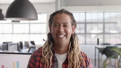 Portrait-of-happy-biracial-businessman-with-dreadlocks-smiling-in-creative-office-in-slow-motion