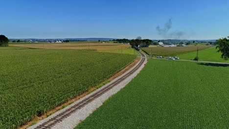 una vista aérea que viaja frente a un antiguo tren de pasajeros de vapor, que sopla humo negro que viaja a través de campos de maíz y ricas tierras de cultivo en un día de verano sin nubes