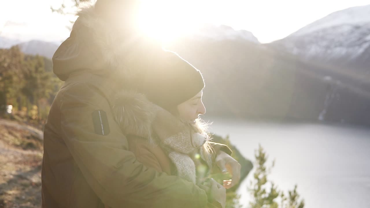 겨울 코트를 입은 행복한 젊은 커플이 포옹하고, 포옹하는 사람들이 호수와 산 정상의 멋진 전망을 바라보고 있습니다. 아름다운 산 자연 풍경을 감상하십시오. 렌즈 플레어