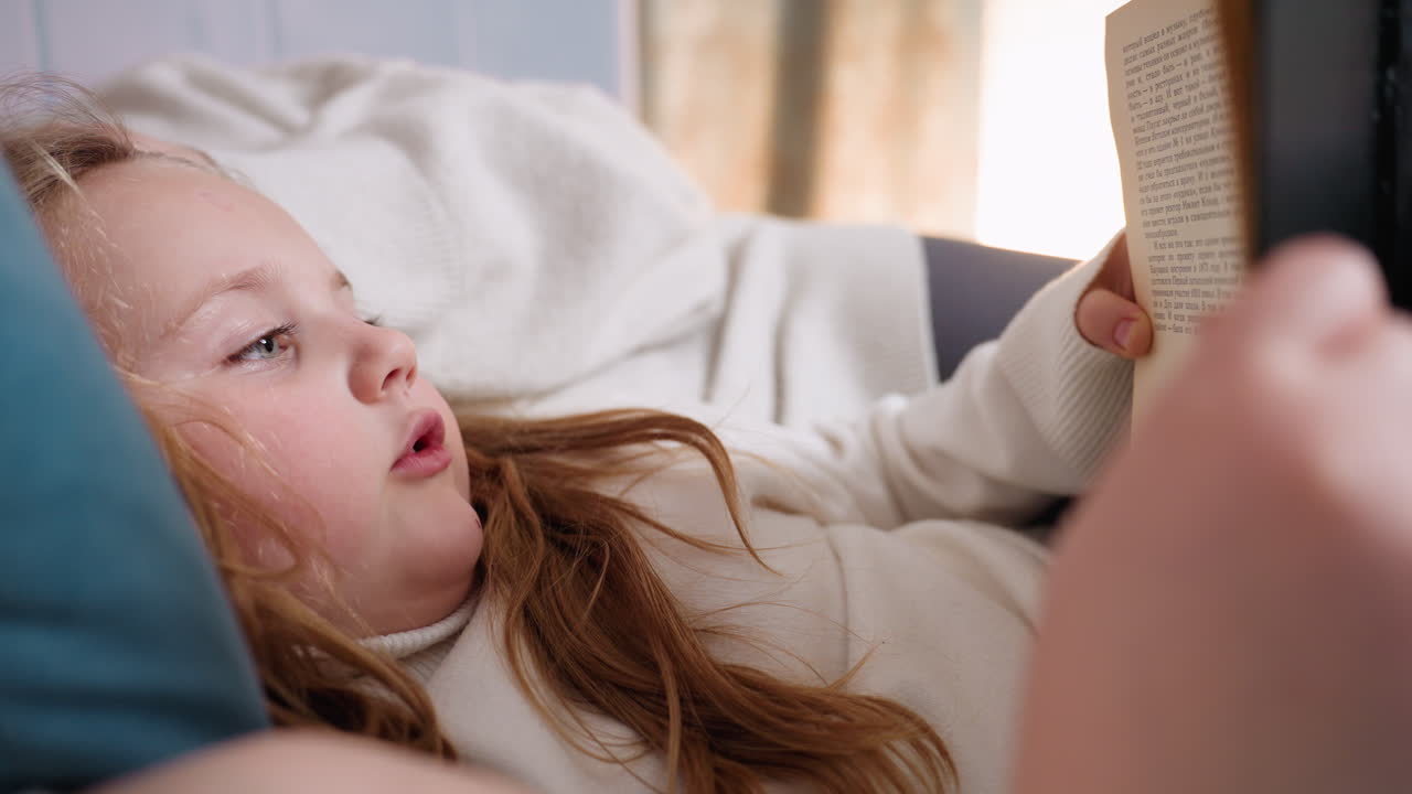 Curious young girl lying on bed holding book reading aloud, focused expression showing engagement with story, cozy atmosphere with warm light and comfortable clothing peaceful educational family moment