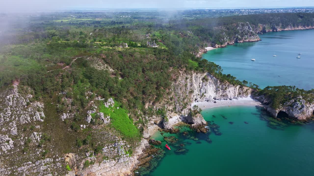 Drone pulling back from Île Vierge beach in Crozon peninsula, showing cliffs, rocks, vegetation, forest, morning mist and a red boat. - Brittany in France