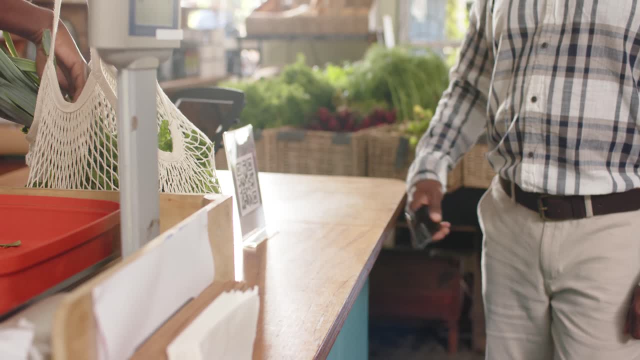 sección media de un hombre afroamericano de alto nivel pagando con un teléfono inteligente en una tienda de alimentos saludables, cámara lenta