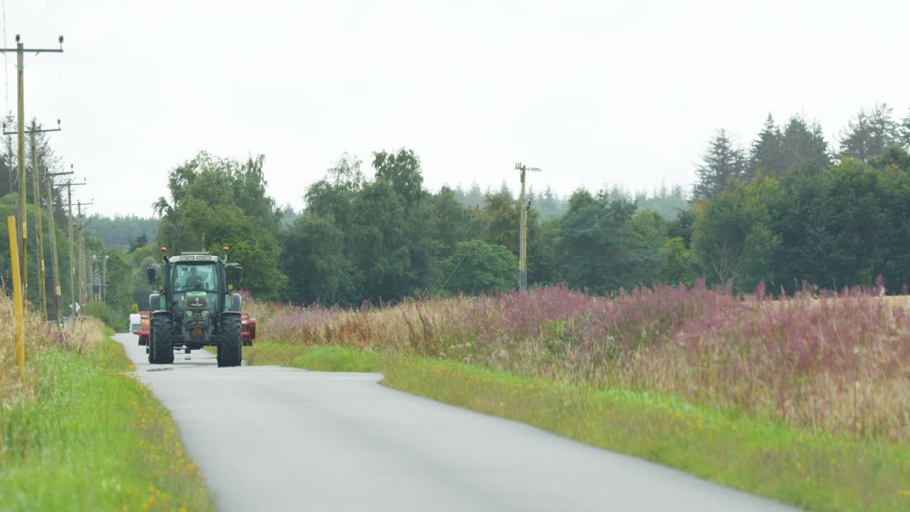A green tractor travels along a narrow country road bordered by wildflowers and fields under overcast daylight, with steady camera framing and natural lighting