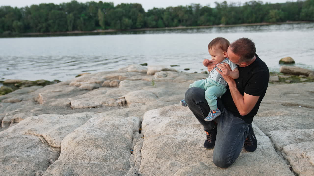Father holding his little baby boy on one knee. Loving daddy kissing his son's cheek and back of the neck. Family concept. River at backdrop.