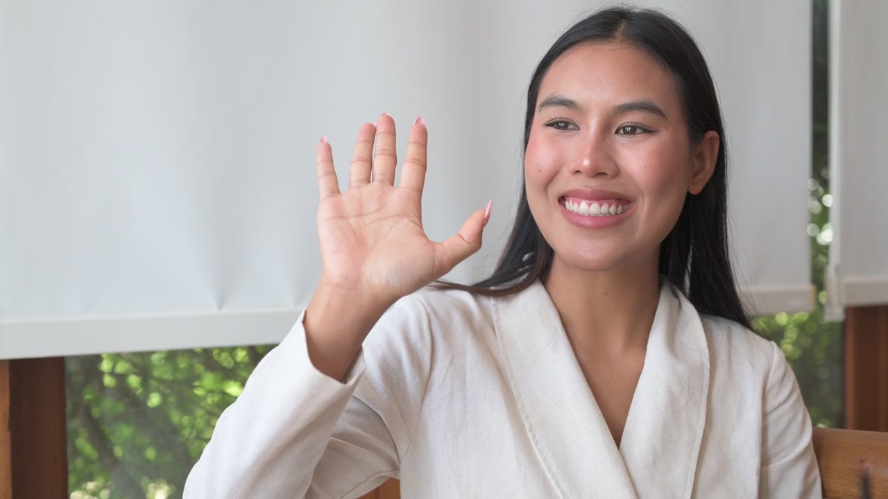 Giovane donna asiatica sorridente che saluta con la mano con un gesto amichevole, guardando altrove