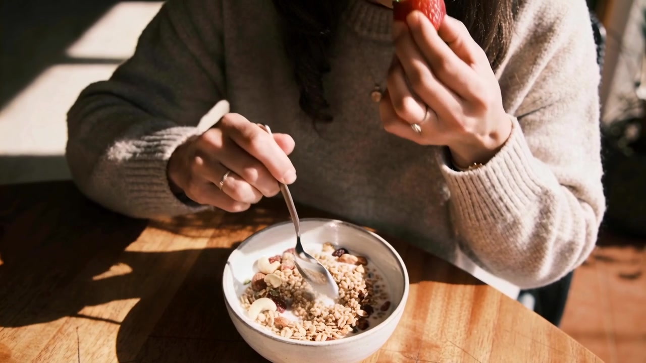 Una donna con un maglione, illuminata dal sole, mescola i cereali da mangiare e porta una fragola alla bocca, seduta a tavola a casa con una ciotola.