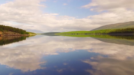 Drone-gliding-forward-1m-over-smooth-surface-lake-on-sunny-day-with-mountains-and-clouds-in-distance