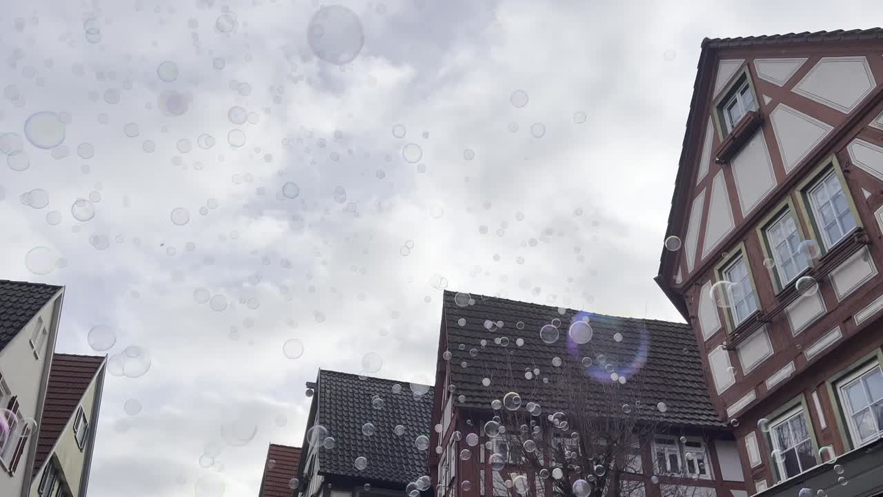 Zeepbellen zweven door een historische straat in Besigheim met traditionele vakwerkhuizen en een bewolkte lucht. Stedelijk toerismebeeld met opwaartse camerahoek en bewegende bellen.