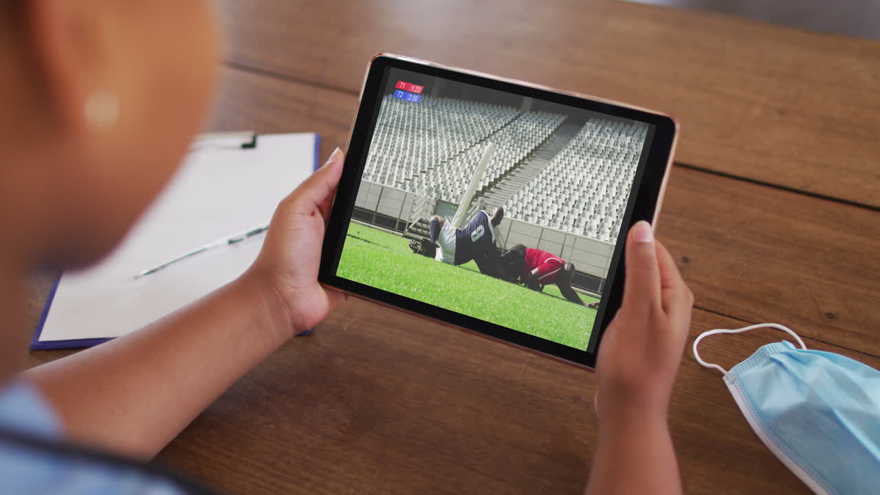 태블릿에서 럭비 경기를 시청하는 얼굴 마스크와 함께 테이블에 앉아있는 여성의 복합