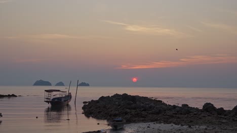 Wide-shot-of-boat-lying-on-shore,-red-sun-setting-on-the-horizon