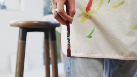 hand of biracial female artist in apron holding paint brush in art studio, slow motion