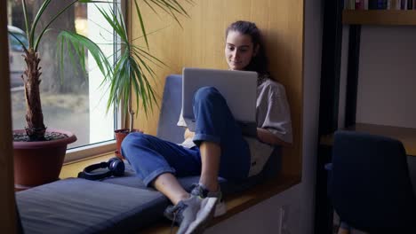 Young-attractive-female-freelancer-sitting-on-windowsill-in-cafe-and-working-on-laptop