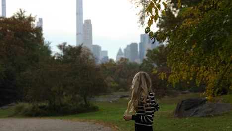 una mujer rubia de pie en el parque central de la ciudad de nueva york con rascacielos en el fondo