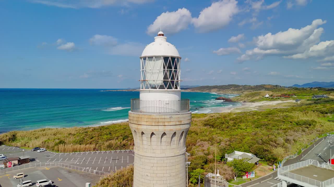 A close-up aerial orbit of the Hinomisaki Lighthouse tower, revealing the stunning turquoise sea, sandy beach, and lush green landscape of the Shimane Peninsula on a bright, sunny day