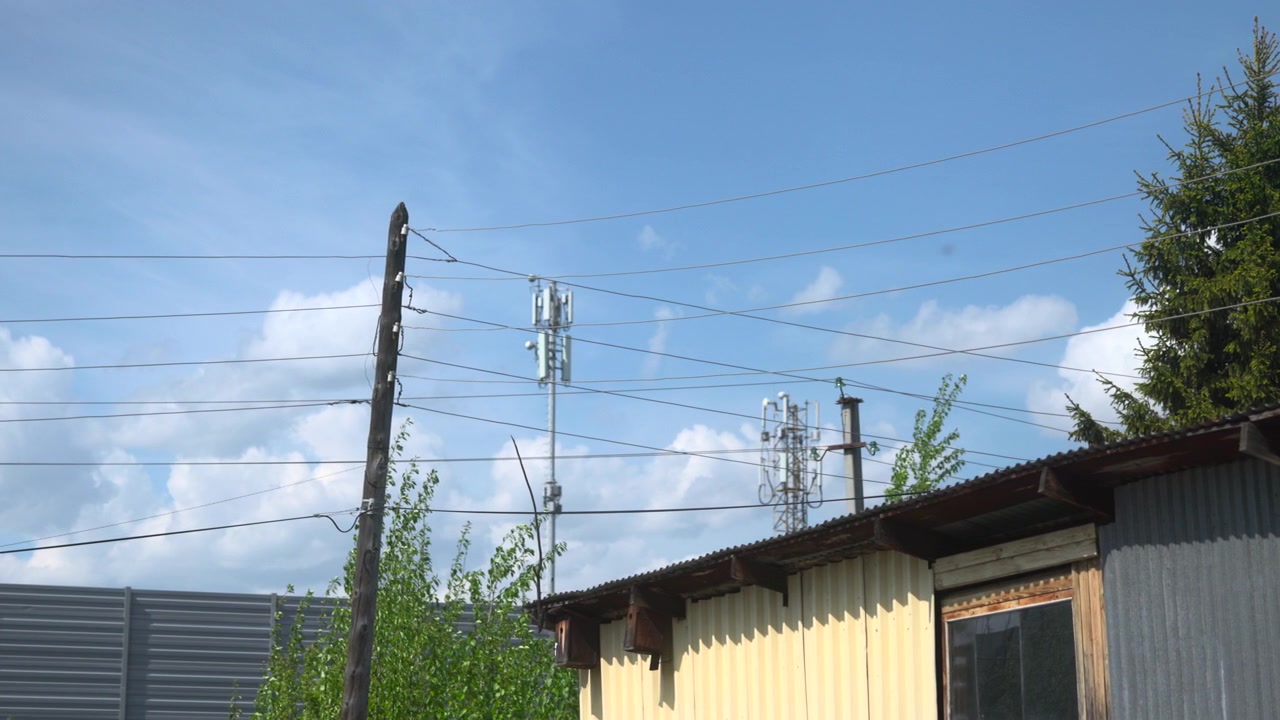 Overhead utility lines and poles rise above low industrial buildings with a telecom tower, trees, and a bright blue sky with scattered clouds in a daytime