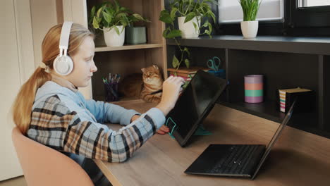 the girl draws, uses a digital graphics tablet. inspiration and new technologies. she is being watched by a red cat sitting on a shelf