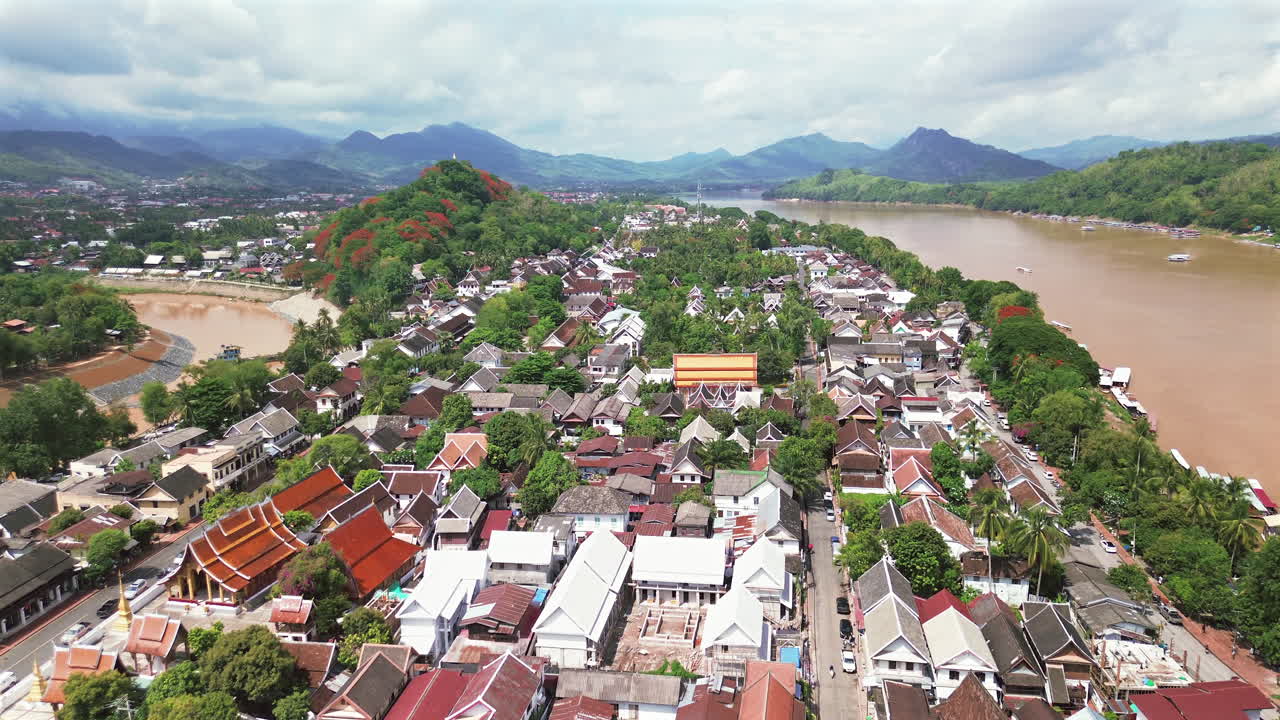 High drone establishing panoramic of Luang Prabang rooftops, river curves, and surrounding mountains, Laos with traditional architecture