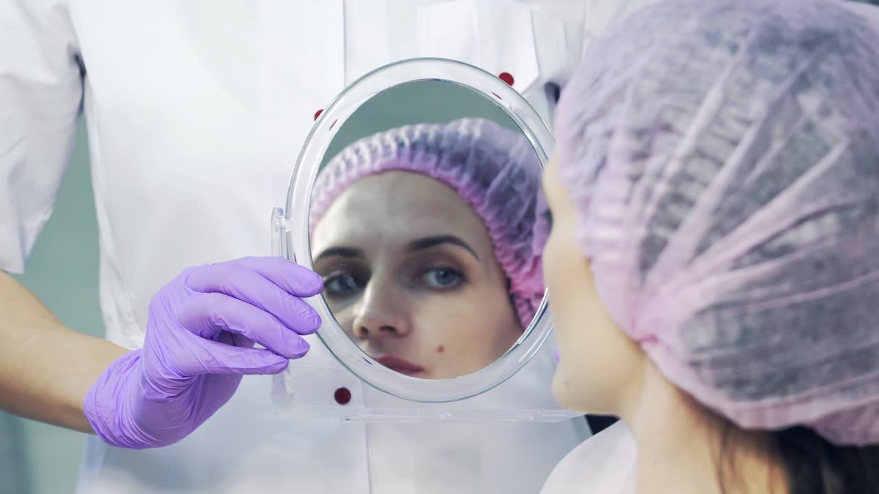 Doctor holds the mirror and the patient looks at her lips and smiles in a beauty salon. The process of lips enhancement