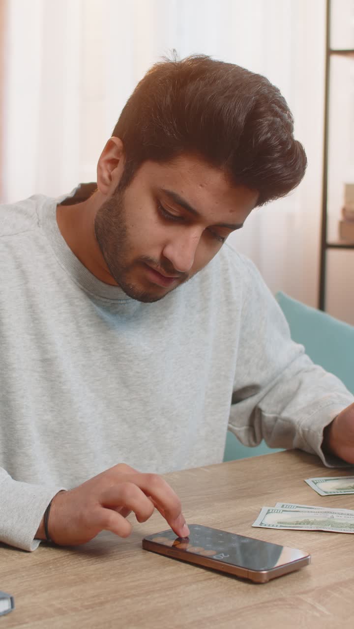 Young indian man planning expenses organizing cash in dollars using smartphone calculator at home