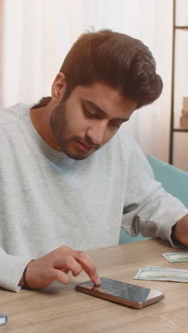 young indian man planning expenses organizing cash in dollars using smartphone calculator at home