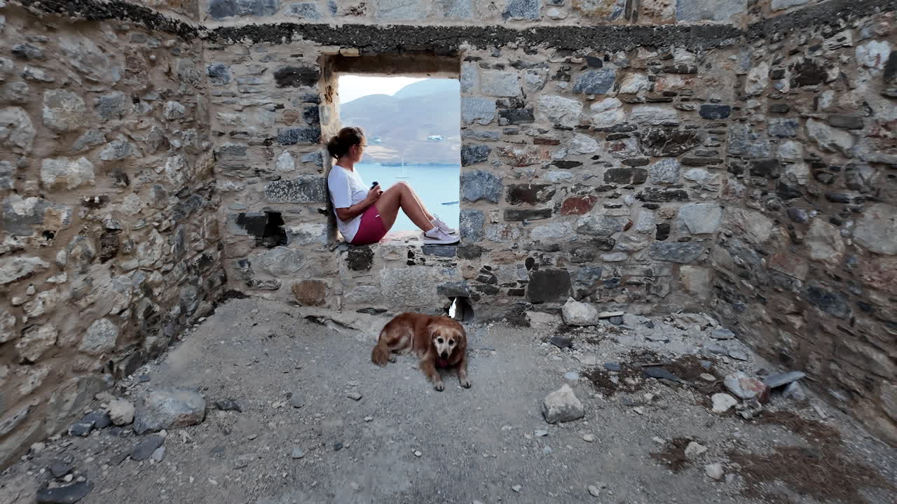 Greece, Astypalea Island, iconic castle wall, a woman sitting at the window sash of the old wall and a golden retriever is lying on the floor.Camera passes the woman and looks over to the Aegean Sea.