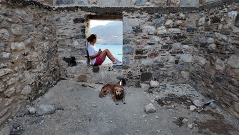 greece, astypalea island, iconic castle wall, a woman sitting at the window sash of the old wall and a golden retriever is lying on the floor.camera passes the woman and looks over to the aegean sea.
