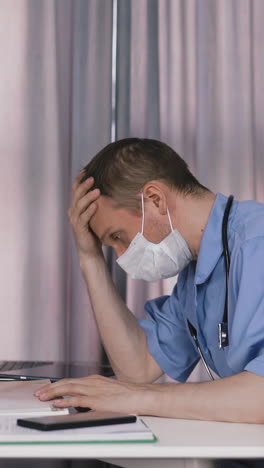 tired skilled doctor wearing blue uniform and protective mask reads patient medical records sitting at table with laptop and removes guise in office