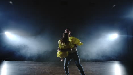 camera is zooming and shooting from the full shot to the medium one in slow moting a young girl in a yellow dress and a man in a white shirt dancing on the stage in spotlights and smoke