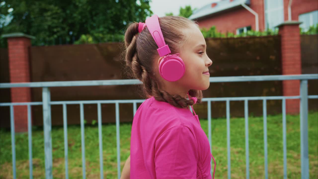 Young Girl Enjoying Music While Walking Outdoors, Wearing Bright Pink Headphones in a Lush Green Environment with a Background of Colorful Houses