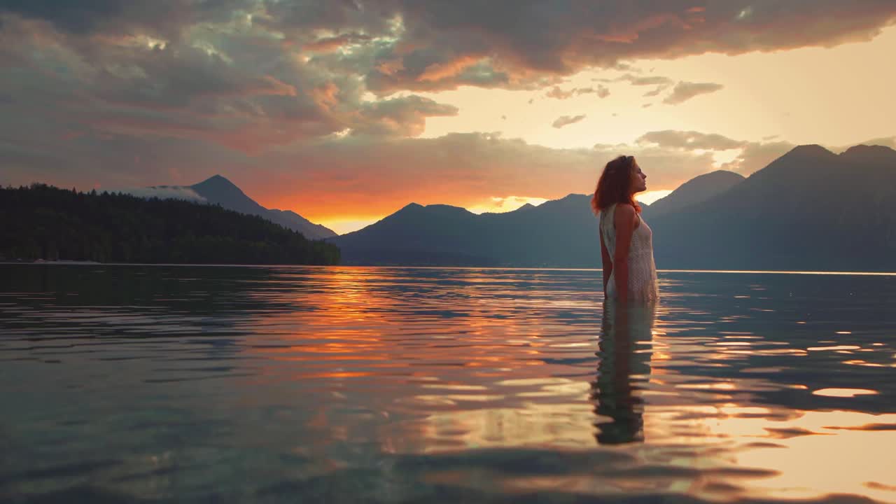 Dreamy Cinemagraph - Seamless video loop of a beautiful young model girl standing in the calm waves of lake Walchensee in Southern Germany-Bavaria in a white fashion dress by sunset, facing the sun.