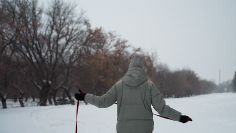 woman in grey winter jacket and knit hat stands alone in snowy park path holding red leash, facing away under overcast sky, surrounded by leafless trees, evoking quiet moment of reflection and solitude