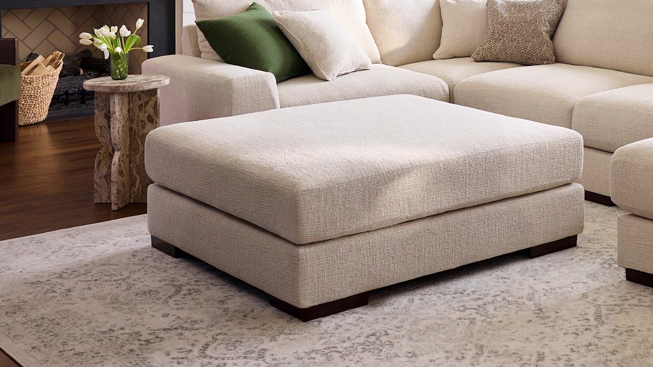 Slow zoom out of a beige upholstered farmhouse ottoman in a professionally staged living room with warm natural light