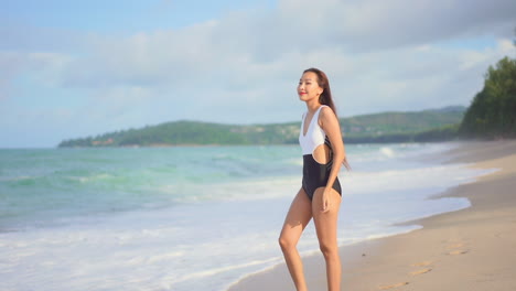 Joven-Asiática-Con-Traje-De-Baño-Blanco-Y-Negro-Caminando-Por-La-Línea-De-Playa-De-La-Pintoresca-Isla,-Siguiendo-El-Tiro,-En-Cámara-Lenta