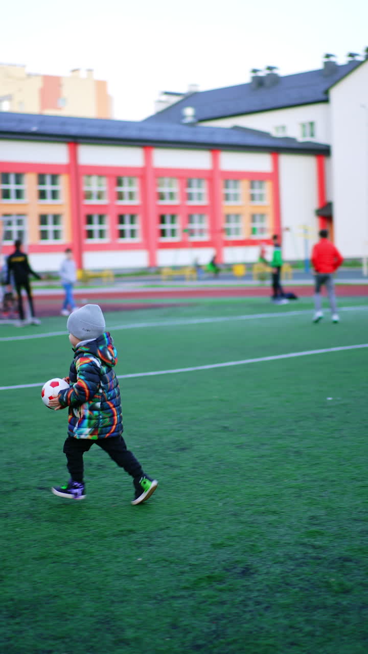 Energetic toddler boy runs carrying a ball in hands. Two-year-old child playing active games outdoors at stadium. Vertical video.