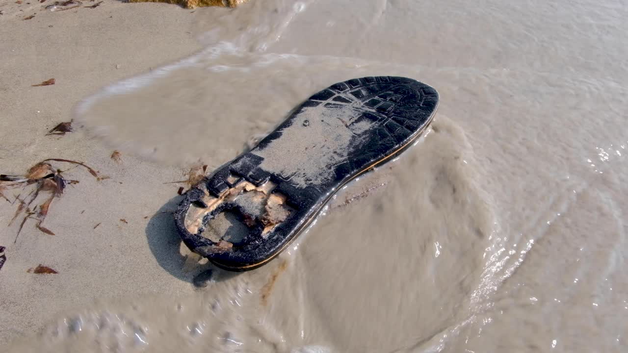 A single lost and partially decomposed black flip flop on a sandy beach with waves washing over it polluting and littering on a remote tropical island