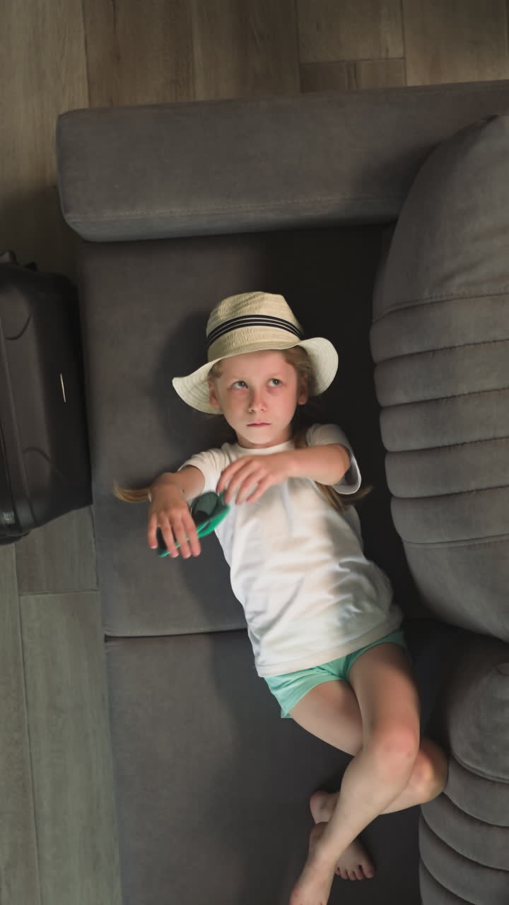 mamá viene a la niña acostada en el sofá y jugando con gafas de sol. preescolar aburrido mueve las manos en el baile listo para el vuelo al país tropical vista superior