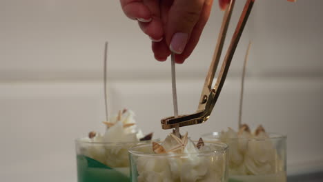pastry chef gently holds straw inside glass cup of decorated ice cream dessert, carefully trims long straw with scissors, focusing on presentation and precision while surrounded by whipped topping