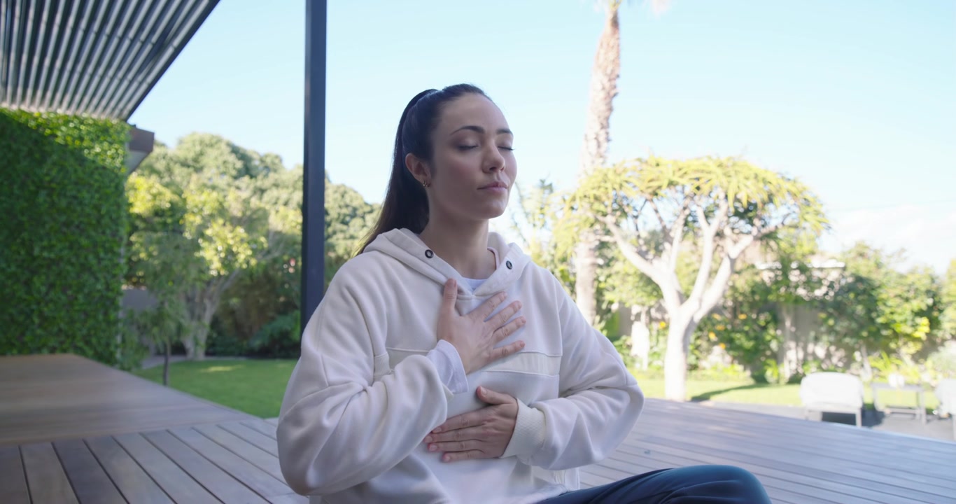 Mujer sentada con las piernas cruzadas en una colchoneta verde, con la mirada hacia adentro, respirando para la calma y con sudadera con capucha.