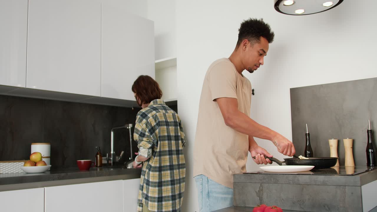 A young man with Black skin color in a beige T-shirt puts ready-made scrambled eggs on a plate while his young adult girlfriend with a bob hairstyle washes the dishes while preparing breakfast in the morning at a modern apartment