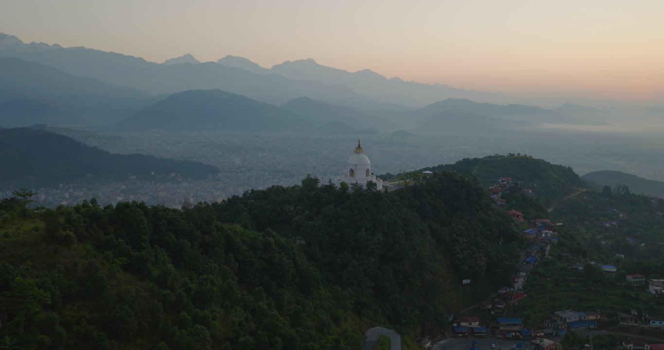 Aerial shot of the locality around World Peace Stupa with an expansive cloudy horizon and surrounding green forest landscape in Pokhara, Nepal.