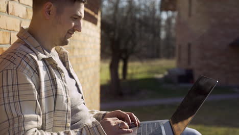 side view of caucasian man using laptop while sitting on a bench outside a country house