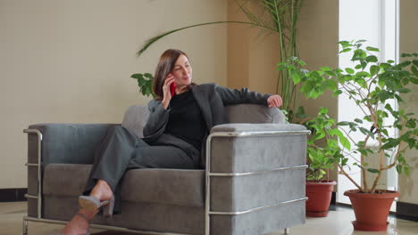 confident businesswoman in formal outfit lounging casually on stylish sofa while talking on smartphone, surrounded by potted plants and natural daylight in modern spacious office waiting area