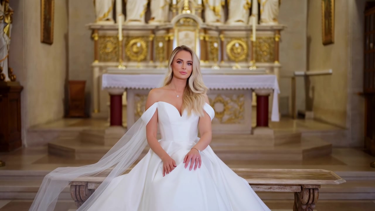 Sposa bionda in abito da sposa bianco seduta in primo piano al centro di una chiesa riccamente decorata vicino all'altare, con luce calda e soffusa e una cerimonia tranquilla