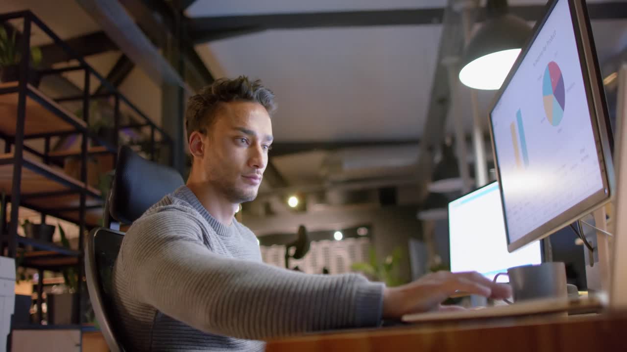 Afslappet forretningsmand af blandet etnisk baggrund, der sidder ved skrivebordet og bruger computere på kontoret om natten, slowmotion.