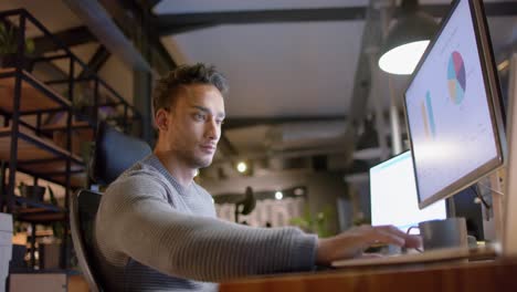 Hombre-De-Negocios-Casual-Birracial-Sentado-En-El-Escritorio-Usando-Computadoras-En-La-Oficina-Por-La-Noche,-Cámara-Lenta