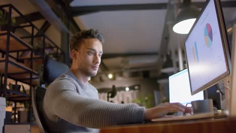 biracial casual businessman sitting at desk using computers in office at night, slow motion