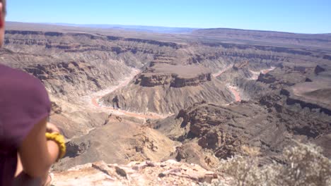 Over-the-Shoulder-of-Young,-Blonde-Millenial-Woman-Sitting-and-looking-towards-the-The-Fish-River-Canyon