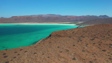 Cinematic-drone-shot-of-Balandra-Beach,-passing-over-the-red-hills,-turquoise-waters-and-white-sand-beaches,-descending-over-the-hills