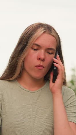 mujer joven caminando al aire libre, sosteniendo el teléfono al oído, mirando involucrado en una llamada telefónica con expresión reflexiva, el fondo presenta vegetación exuberante y árboles